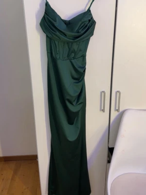 Grön långklänning med slits - Elegant grön långklänning med smala axelband och draperad urringning. Klänningen har en snygg slits på sidan och figurnära passform som framhäver siluetten. Perfekt för speciella tillfällen där du vill sticka ut.