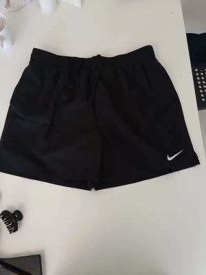 Svarta träningsshorts från Nike - Snygga svarta träningsshorts från Nike med klassisk vit logga på benet. Shortsen har elastisk midja med snörning för skön passform och är tillverkade i ett lätt och funktionellt material som passar perfekt till träning.
