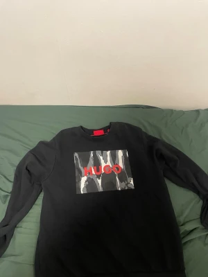 Svart sweatshirt från Hugo - Cool svart sweatshirt från Hugo med ett stort tryck på bröstet i grått, vitt och rött där det står HUGO. Tröjan har rund halsringning och långa ärmar. Perfekt för en avslappnad och trendig stil.