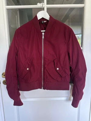 Vinröd bomberjacka - Klassisk vinröd/burgundy bomberjacka från Divided med silvrig dragkedja och två fickor framtill med tryckknappar. Jackan har ribbade muddar vid krage, ärmslut och nederkant. Lätt vadderad med fint lyster i tyget! 💫 