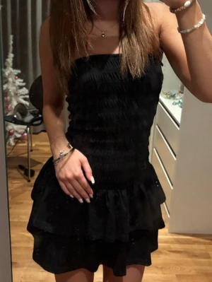 Svart axelbandslös volangklänning - Säljer en svart axelbandslös klänning med smockad överdel och volangkjol. Klänningen har en kort längd och sitter tight upptill, perfekt för dig som gillar en feminin och trendig look. Passar till många olika tillfällen!