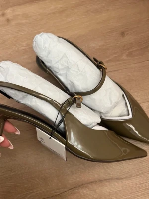 Olivgröna lackpumps från Zara - Snygga olivgröna pumps från Zara i glansigt lackmaterial. Skorna har spetsig tå, smal klack och en elegant rem med guldfärgat spänne runt ankeln. Perfekta för dig som vill ha en trendig och stilren look.