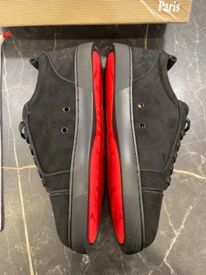 Svarta sneakers från Christian Louboutin - Säljer ett par svarta sneakers från Christian Louboutin med ikoniska röda sulor och diskret logga undertill. Skorna är tillverkade i mocka och har svarta snören samt metallhål för snörning. Perfekta för dig som vill ha en lyxig och stilren look.
