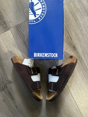 Bruna Birkenstock Arizona sandaler - Säljer ett par klassiska Birkenstock Arizona sandaler i mörkbrunt mocka🤗 Sandalerna är i fint skick, hör av er vid frågor🙌🏻