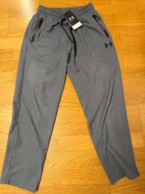 Grå mjukisbyxor från Under Armour - Snygga grå mjukisbyxor från Under Armour med resår i midjan och svarta dragsnören. Byxorna har två fickor med dragkedja och logga på vänster ben. Perfekta för chill eller träning, tillverkade i mjukt och bekvämt material. Aldrig använda och den kommer med tag.