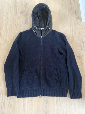 Gran Sasso cardigan - Snygg mörkblå gran sasso cardigan med dragkedja framtill och två stora fickor. Huva i kontrasterande material med dragsko. Stickad känsla i tyget och ribbade muddar vid ärmslut och nederkant. Perfekt för en avslappnad och cool stil. Köpt på Ströms för 4499 | Skick: 8/10 | Storlek: 52/L | Meddela vid eventuella frågor eller funderingar! 