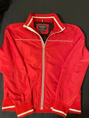 Röd vindjacka från East West - Snygg röd vindjacka från East West med vita detaljer och dragkedja framtill. Jackan har ribbade muddar och krage med vita ränder, samt en sportig look. Tillverkad i lätt och slitstarkt syntetmaterial, perfekt för blåsiga dagar.