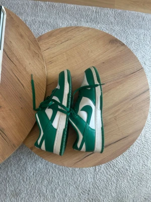 Nike Dunk Low gröna och vita sneakers - Säljer ett par Nike Dunk Low sneakers i grönt och vitt. Skorna har klassisk låg siluett, gröna snören och sula samt vit bas med gröna detaljer och swoosh. Perfekta för dig som vill ha en clean och sportig look.