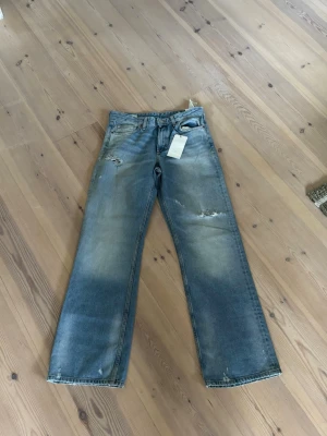 Blå raka jeans från Zara - Säljer ett par blå raka jeans från Zara med slitna detaljer och ljus tvätt. Jeansen har fem fickor, normal midja och klassisk knappgylf. Materialet är bomull och de har en avslappnad passform som ger en cool och trendig look. Små så passar 38
