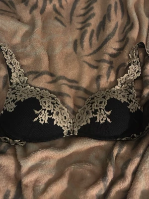 Svart bh med beige spetsdetaljer från intimissimi - Snygg svart bh med vadderade kupor och dekorativa beige spetsdetaljer längs kanterna och axelbanden. Bh:n har en klassisk bygelmodell och tunna axelband med spets. Perfekt för dig som gillar en elegant och feminin stil.