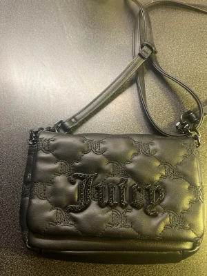 Svart Juicy Couture axelväska - Snygg svart axelväska från Juicy Couture med quiltat mönster och broderade JC-loggor. Väskan har justerbar axelrem och ett stort Juicy-tryck på framsidan. Insidan är fodrad i ljusrosa tyg med Juicy Couture-tryck. Perfekt för att lyfta din outfit.