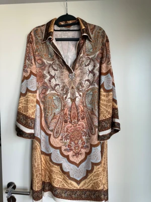 Mönstrad kortklänning i bruna toner - Unik kortklänning med bohemiskt mönster i beige, brunt och inslag av blått. Klänningen har krage, v-ringning och trekvartsärm. Tillverkad i ett mjukt, lätt material viskos som känns lyxigt mot huden. Perfekt för dig som gillar statement-plagg med coola detaljer.