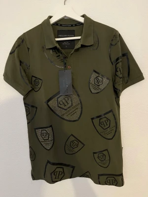 Olivgrön Philipp Plein pikétröja - Snygg olivgrön pikétröja från Philipp Plein med svart logomönster och detaljer i strass. Klassisk krage, korta ärmar och knappslå framtill. Tillverkad i bomull för en bekväm känsla. Perfekt för dig som gillar streetwear med exklusiv touch.