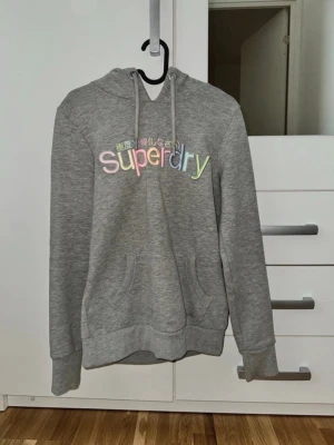 Grå Superdry hoodie med regnbågstryck - Säljer en grå hoodie från Superdry med broderad logga i regnbågsfärger och japansk text på bröstet. Hoodien har huva med dragsko, magficka och ribbade muddar. Perfekt för dig som gillar en avslappnad och färgglad stil.