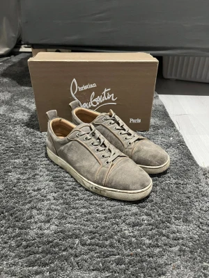 Gråa Christian Louboutin sneakers - Snygga gråa sneakers från Christian Louboutin i mocka med klassisk röd sula och lågt skaft. Skorna har vita sulor, gråa skosnören och diskreta sömdetaljer. Perfekta för dig som vill ha något exklusivt och stilrent.