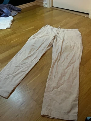 Beiga linnebyxor från Uniqlo - Säljer ett par beiga linnebyxor från Uniqlo med rak passform och resår i midjan. Byxorna är tillverkade i en skön blandning av linne och bomull, perfekta för en avslappnad stil. Midjan är justerbar med snörning för extra komfort.