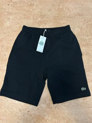 Svarta shorts från Lacoste - Svarta shorts från Lacoste med elastisk midja och klassisk krokodil-logga broderad vid benet. Regular fit och tillverkade i mjuk bomull. Köpta från zalando 2a maj för 900kr. Aldrig använda. Kan skicka med kvitto i chatten 