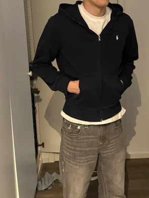 Mörkblå hoodie från Polo Ralph Lauren - Klassisk mörkblå hoodie från Polo Ralph Lauren med dragkedja framtill, två fickor och justerbar huva med snören. Diskret logga broderad i vitt på bröstet. Tillverkad i mjuk bomullsblandning för en skön och avslappnad look.