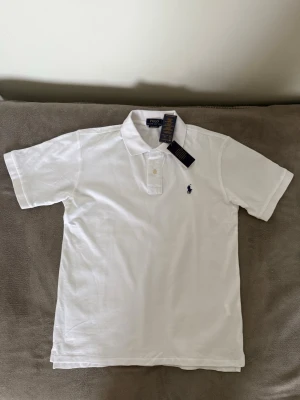  Polo Ralph Lauren Piketröja - Klassisk vit pikétröja från Polo Ralph Lauren med korta ärmar och broderad logga på bröstet. Tröjan har krage, knappar framtill och är tillverkad i mjuk bomull. Passformen är slim och stilen är tidlös och clean.