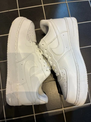 Nike Air Force 1 vita låga sneakers - Klassiska Nike Air Force 1 sneakers i helvitt läder med perforerad tåbox, vita skosnören och den ikoniska swoosh-loggan på sidorna. Tjock sula och låg profil ger en clean och tidlös look som alltid funkar. Perfekta för dig som gillar streetwear och vill ha något stilrent. KOLLA IGENOM ALLA BILDER ÄR NI SNÄLLA!🙏 Har använt dem en gång!