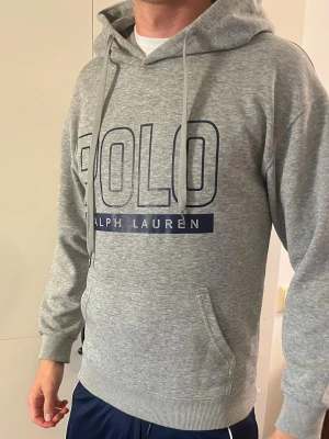 Grå hoodie från Polo Ralph Lauren - Snygg grå hoodie från Polo Ralph Lauren med stor logga i mörkblått framtill. Klassisk känguruficka, ribbade muddar och justerbar huva med dragsko. Tillverkad i mjukt sweatshirtmaterial som är skönt mot huden. Perfekt för en avslappnad och trendig look.