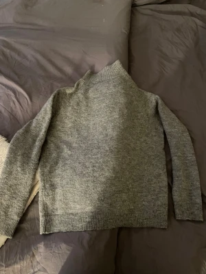 Grå stickad polotröja från Envii - Mysig grå stickad polotröja från Envii med ribbade muddar och hög krage. Tröjan har en enkel, rak passform och är perfekt för kyliga dagar. 