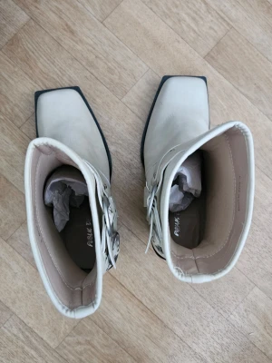Vita cowboy boots med silverdetaljer - Snygga vita cowboy boots från Public Desire med fyrkantig tå, bred klack och coola silverfärgade spännen på sidan. Boots i konstläder med högt skaft och trendig design som sticker ut. Har aldrig använt. Det finns två sträck (se bilden). Går nog att ta bort med tvål och vatten ellee med Isopropylalkohol (ej aceton!). 
