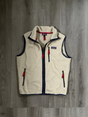 Beige fleeceväst från Patagonia - Säljer en beige fleeceväst från Patagonia med marinblå detaljer och röda dragkedjor. Västen har tre fickor med dragkedja och stårkrage. Perfekt för lager-på-lager och outdoor-vibe.