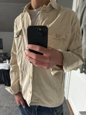 Overshirt  - Säljer en beige overshirt i manchester med klassisk krage och bröstfickor med knappar. Skjortan har långa ärmar och en avslappnad passform, perfekt att bära öppen över en t-shirt för en casual look.