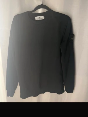 Svart sweatshirt från Stone Island - Svart långärmad sweatshirt från Stone Island med klassisk rund hals och ikonisk patch med kompasslogga på vänster ärm. Tröjan har en clean look och är tillverkad i mjukt bomullsmaterial. Perfekt för dig som gillar streetwear och stilrena plagg.