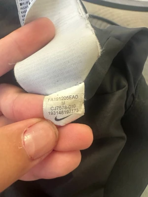 Svart vindjacka från Nike - Snygg svart vindjacka från Nike i storlek M. Jackan har en klassisk design med huva och dragkedja, samt små Nike-loggor längs insidan. Tillverkad i lätt och slitstarkt material som passar perfekt för blåsiga dagar.