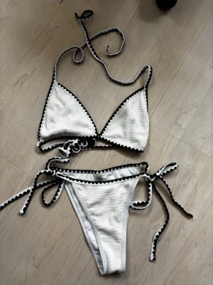 Vit bikini med svarta detaljer - Snygg vit bikini med svarta sicksack-detaljer längs kanterna. Klassisk trekantsmodell med knyt både i ryggen och nacken samt på sidorna av bikinitrosan. Materialet har en lätt struktur som ger en cool look. Perfekt för stranden eller poolen!