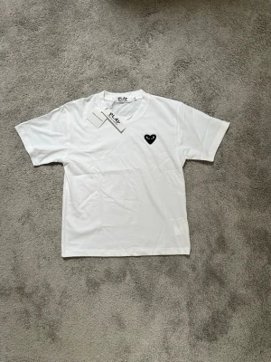 Vit Comme des Garçons PLAY t-shirt - Vit t-shirt från Comme des Garçons PLAY med det ikoniska svarta hjärte-logotypen broderad på bröstet. Klassisk rund halsringning och korta ärmar. Tillverkad i mjuk bomull för en skön och stilren look.