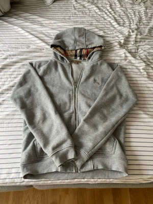 Grå zip hoodie från Burberry - Snygg grå zip hoodie från Burberry, fraktas inom 24 timmar efter köp📦.                                                                                            Storlek: M men är lite kortare i längd.                                                  Snöret har jag tagit bort så det finns ej något snöre 