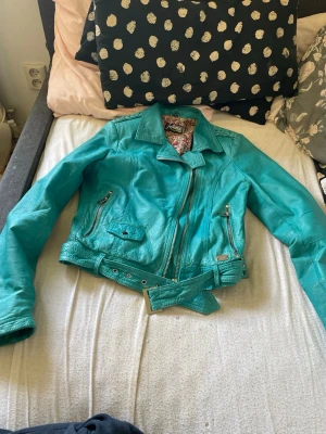 Turkos skinnjacka från Jofama - Snygg turkos skinnjacka från Jofama med klassisk bikerstil, sned dragkedja, två framfickor med dragkedja och bälte i midjan. Insidan har ett blommigt foder i rosa, vitt och svart. Perfekt för dig som vill sticka ut med färg.köpt för 2000