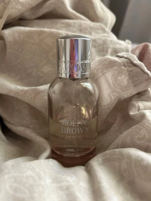 Molton Brown Suede Orris Hair Mist - Halvfull elegant hårmist från Molton Brown i doften Suede Orris. Flaskan är genomskinlig med rund form och har en stilren silverfärgad spraykork. Perfekt för dig som vill ge håret en lyxig och fräsch doft med en exklusiv touch.