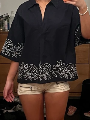 Svart blus med vit brodyr från COS - Stilren svart blus med vit blommig brodyr längs nederkant och ärmar. Blusen har en avslappnad passform, krage och v-ringad hals. Tillverkad i ett vävt, lätt material som känns luftigt. Perfekt för en trendig och avslappnad look. 🥂💗
