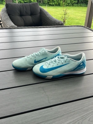 Nike ljusblå fotbollsskor - Snygga ljusblå fotbollsskor från Nike med klassisk blå swoosh på sidan. Skorna har snörning och en låg profil som ger bra bollkontroll. Tillverkade i syntetmaterial för lätt känsla och bra grepp på planen. Perfekta för dig som vill sticka ut på fotbollsplanen.