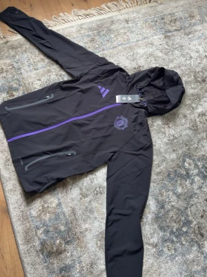Svart Manchester United vindjacka Adidas - Snygg svart vindjacka från Adidas med Manchester United-logga i lila på bröstet och matchande lila dragkedja. Jackan har huva, två sidofickor med dragkedja och justerbar dragsko. Perfekt för dig som vill ha sportig stil och visa ditt lagstöd. Jätte fin och perfekt inför sommaren 
