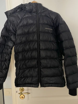Svart dunjacka Peak Performance - Säljer en svart dunjacka från Peak Performance med huva och dragkedja framtill. Jackan har en puffig design med horisontella sömmar och diskret logga på bröstet. Perfekt för kalla vinterdagar och riktigt skön att bära. Dok lite sönder på fram o back sidan men går att sy o fixa 