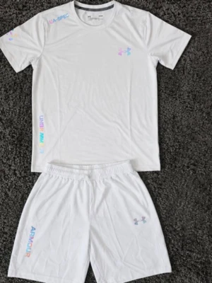 Vitt träningsset från Under Armour - Säljer ett vitt träningsset från Under Armour med t-shirt och shorts. Setet har färgglada loggor och textdetaljer på både tröjan och shortsen. Materialet är lätt och andas, perfekt för träning eller sportaktiviteter.