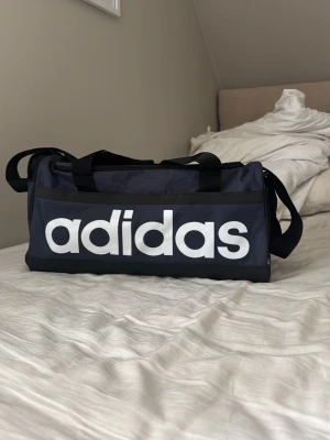 Svart sportbag från Adidas - Snygg svart sportbag från Adidas med stort vitt logotryck på sidan. Väskan har dubbla handtag och justerbar axelrem. Tillverkad i slitstark textil och har en rektangulär form som rymmer mycket. Perfekt för gymmet eller träningen. Aldrig använd