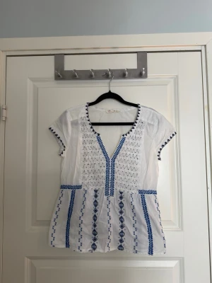 Vit odd molly blus med blå broderier - Söt vit odd molly blus med blå broderade detaljer och korta ärmar. Blusen har v-ringning och ett luftigt fall med dekorativa mönster längs fram- och baksida. Perfekt till sommaren för en fräsch och avslappnad look. Blusen påminner väldigt om mammas Mia och den grekiska sommaren💛
