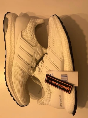 Vita Adidas Ultraboost sneakers - Snygga vita Adidas Ultraboost sneakers med stickad ovandel och klassiska tre ränder på sidan. Skorna har en bekväm Boost-sula och svart yttersula från Continental. Perfekta för dig som gillar stilrena och sportiga sneakers. Skriv för mer info