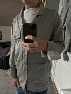 Ljusgrå overshirt - Säljer en stilren ljusgrå overshirt med klassisk krage och tre stora fickor framtill. Overshirten har avslappnad passform och knäpps med knappar. Perfekt att bära över en t-shirt för en casual look.