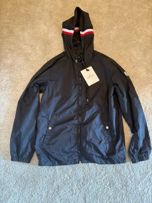 Svart vindjacka från Moncler - Snygg svart vindjacka från Moncler med huva och dragkedja framtill. Jackan har röda och vita ränder på huvan, två fickor med tryckknappar och Moncler-logga på ärmen. Tillverkad i lätt, vattenavvisande materia.
