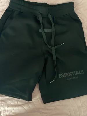 Svarta shorts från Fear of God Essentials - Svarta shorts från Fear of God Essentials med bred resår i midjan och långa snören framtill. Tryckt logga på benet och en extra logodetalj på midjan. Perfekta för en avslappnad och trendig streetwear-look.