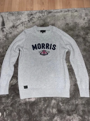 Ljusgrå stickad tröja från Morris - Ljusgrå stickad tröja från Morris med mörkblå text och ett broderat emblem med brittisk flagga på bröstet. Tröjan har rund halsringning, ribbade muddar och nederkant. Perfekt för dig som gillar klassisk och stilren design. Tröjan är storlek S men sitter som XS!
