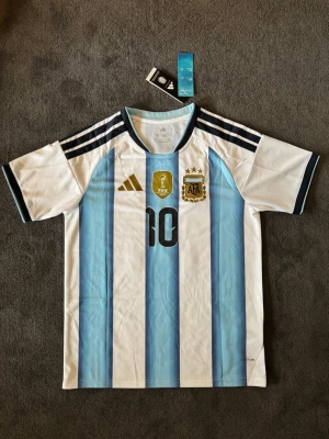 Argentina VM 26/27 hemma Messi 10 - Fotbollströja med Messi på ryggen. Tröjan är helt ny med tagg kvar och i perfekt skick. Perfekt för dig som är supporter, samlar på fotbollströjor eller vill ha en snygg tröja till träning och vardag.                                                                      ⚪ Skickas inom 24 timmar.                                         Vid intresse eller frågor är det bara att skicka 👍
