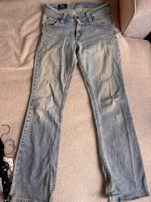 Ljusblå bootcut jeans från Lee - Säljer ett par ljusblå bootcut jeans från Lee med klassisk femficksmodell och slitna detaljer. Har ett mindre hål vid skrevet som lätt går att sy ihop. Det finns ingen storlek men passar en XS som är 160-170 lång.